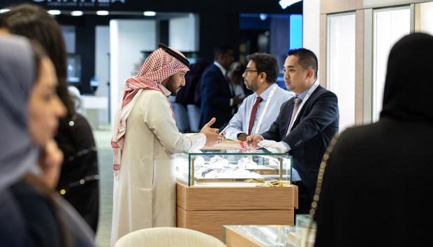 A man in traditional attire talks to two men in suits at a jewellery show. Other people are visible in the background.