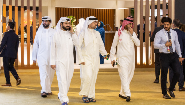 Several men in traditional Middle Eastern attire walk together in an indoor event, with others in Western business attire nearby.